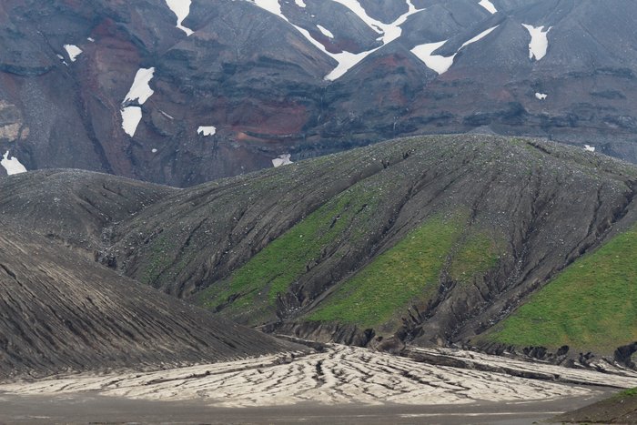 Rills and gullies in an alluvial fan and volcanic cone, in front of a caldera wall exposing lava flows striped by waterfalls.