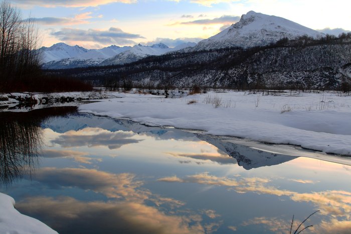 A small channel of the Matanuska River reflects yellow-tinged clouds and the peaks of the Chugach Range.